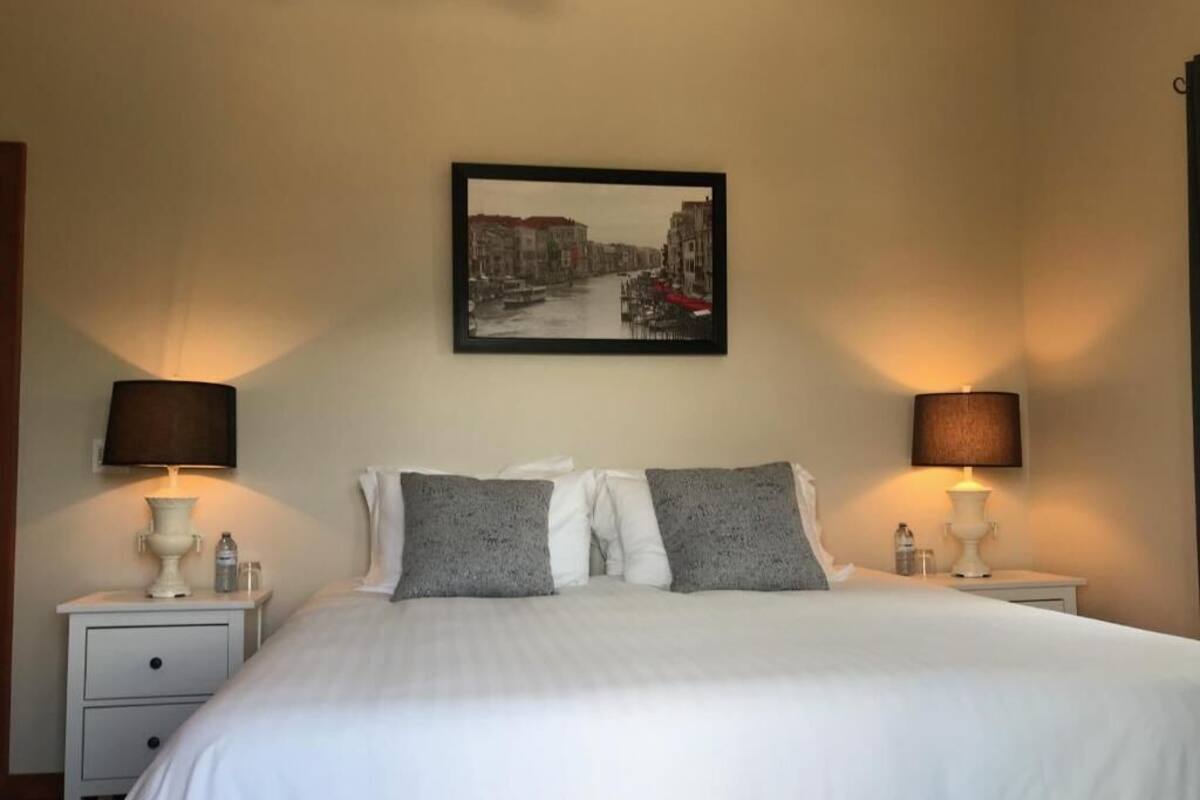 A neatly made bed with white linens and two pillows, flanked by bedside tables with lamps and a framed picture above.