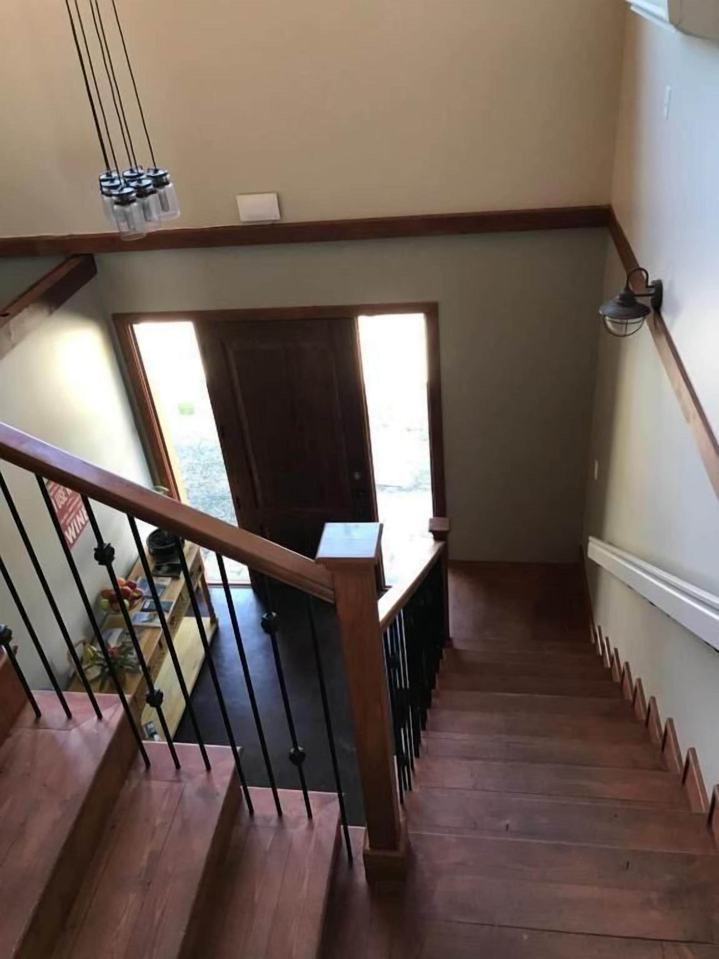 Un escalier en bois avec une rampe, une vue sur l’extérieur par une porte et un luminaire fixé au mur.
