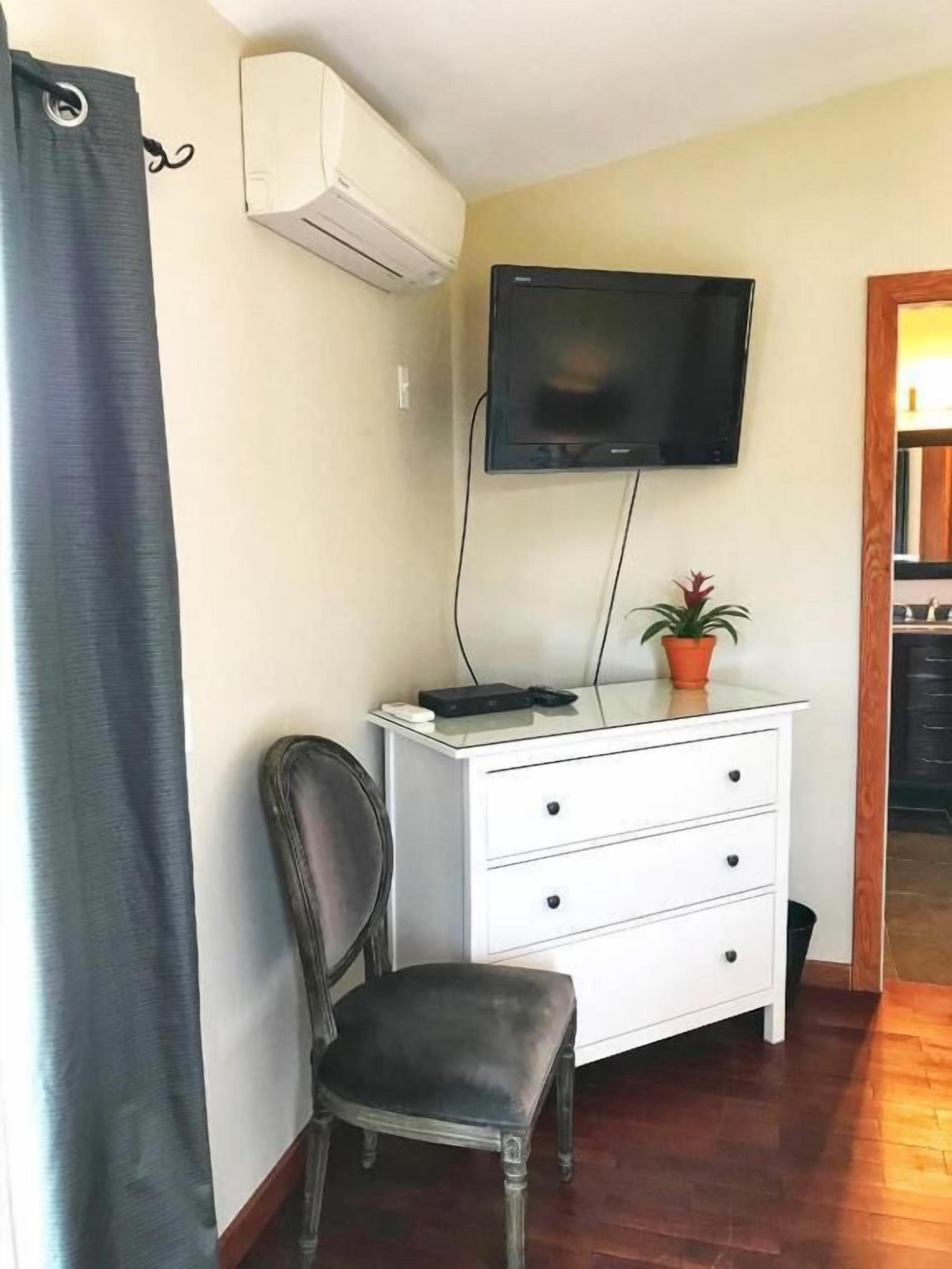 Una habitación con un tocador blanco, un televisor montado en la pared, una planta en maceta y una silla.