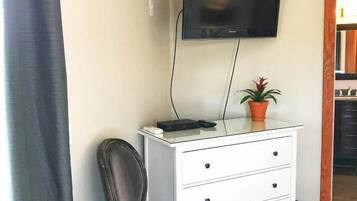 Una habitación con un tocador blanco, un televisor montado en la pared, una planta en maceta y una silla.