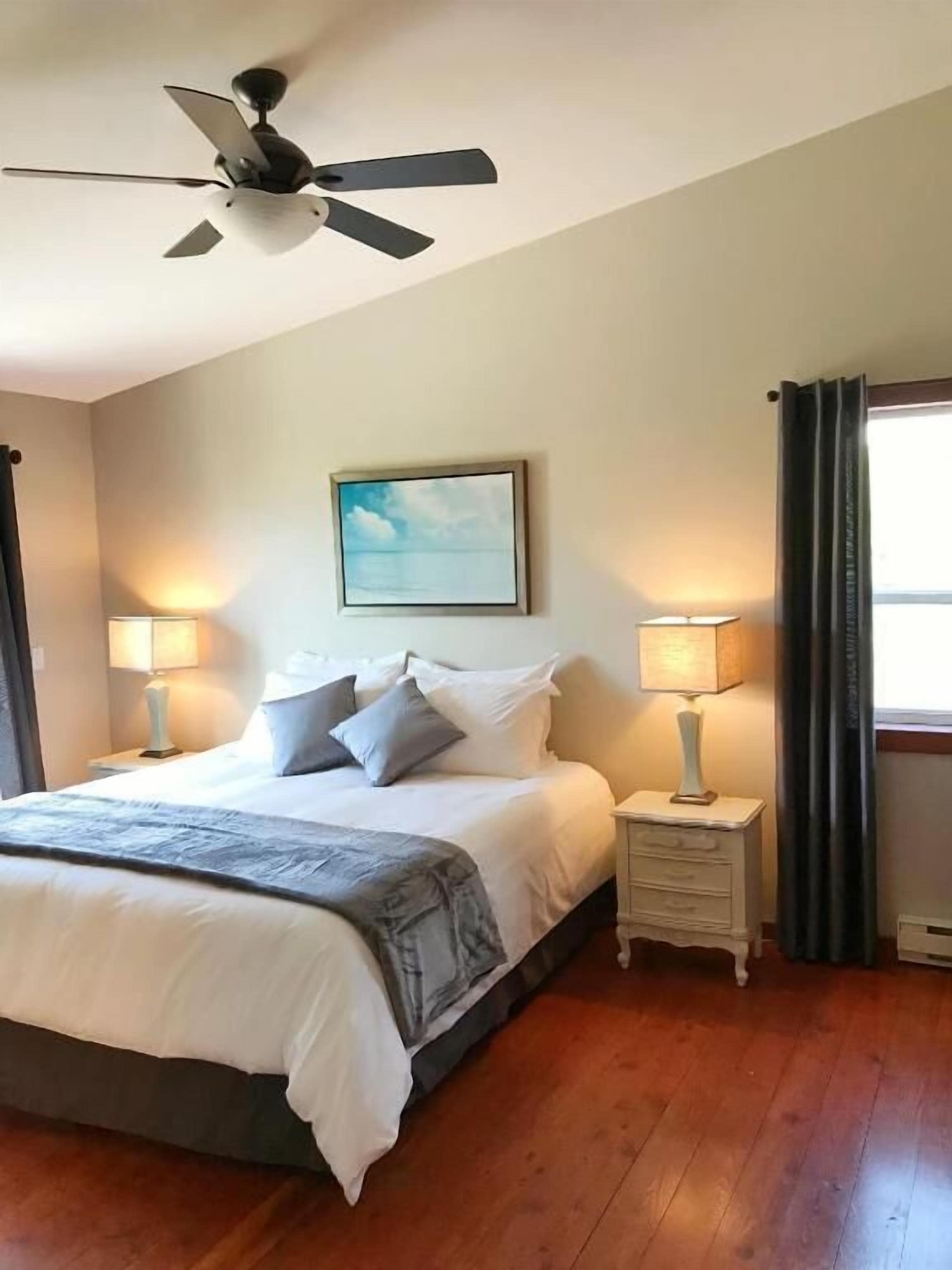 Une chambre à coucher avec un lit, deux tables de chevet avec des lampes, un ventilateur de plafond, un tableau encadré au mur et une fenêtre avec des rideaux.