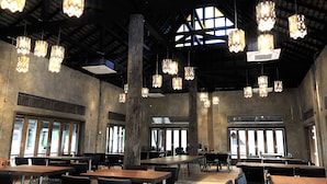 Un vaste espace dédié à la salle à manger, doté de hauts plafonds, de luminaires suspendus et de grandes fenêtres qui laissent entrer la lumière naturelle. Des tables et des chaises y sont disposées pour les repas.