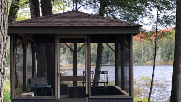 Un pabellón con techo de madera y paredes de cristal, con una mesa y sillas, frente a un fondo de árboles y un lago.
