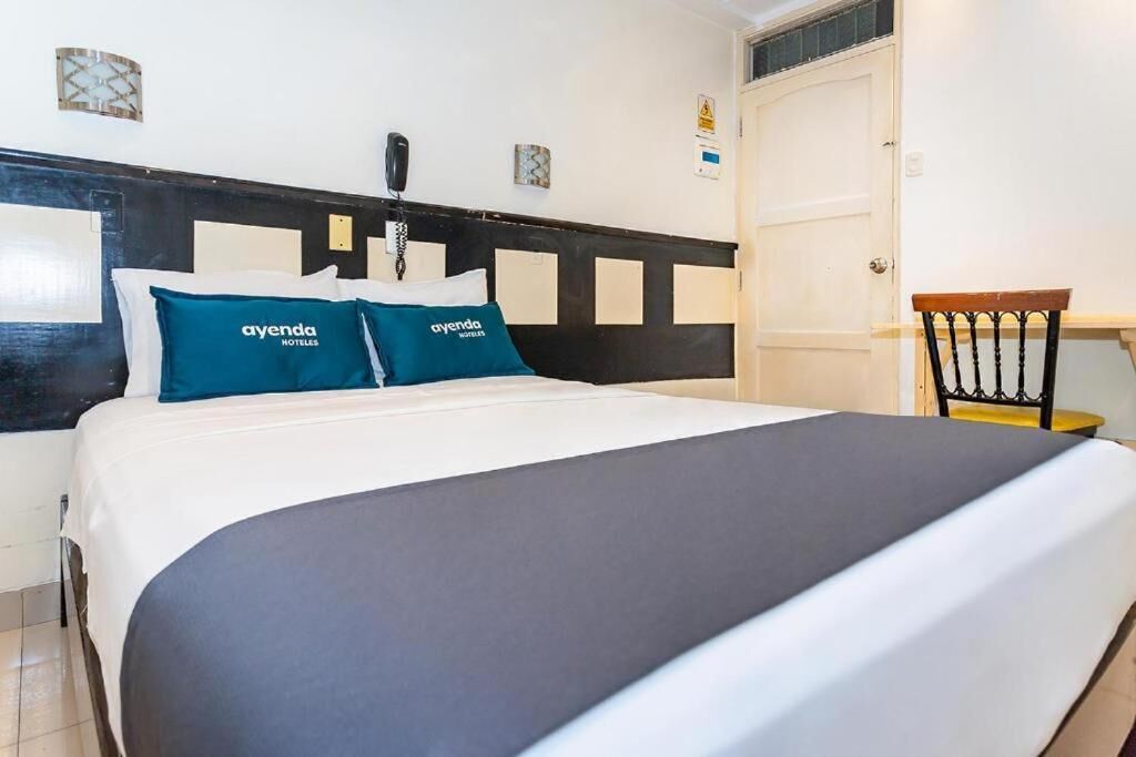 Habitación de hotel con cama, dos almohadas con el nombre del hotel, teléfono en la mesita de noche, una silla y una puerta.