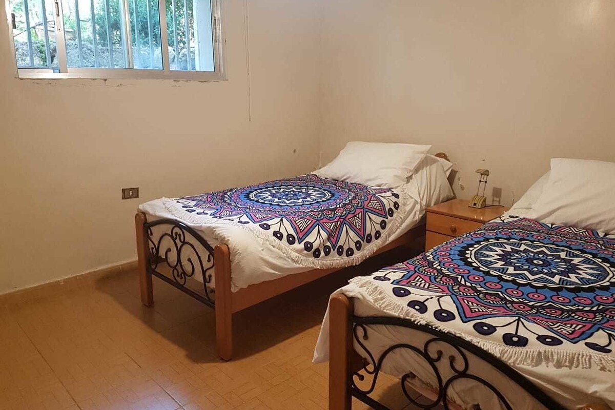 A bedroom with two single beds, each with a patterned bedspread, a window with bars, and a wooden floor.