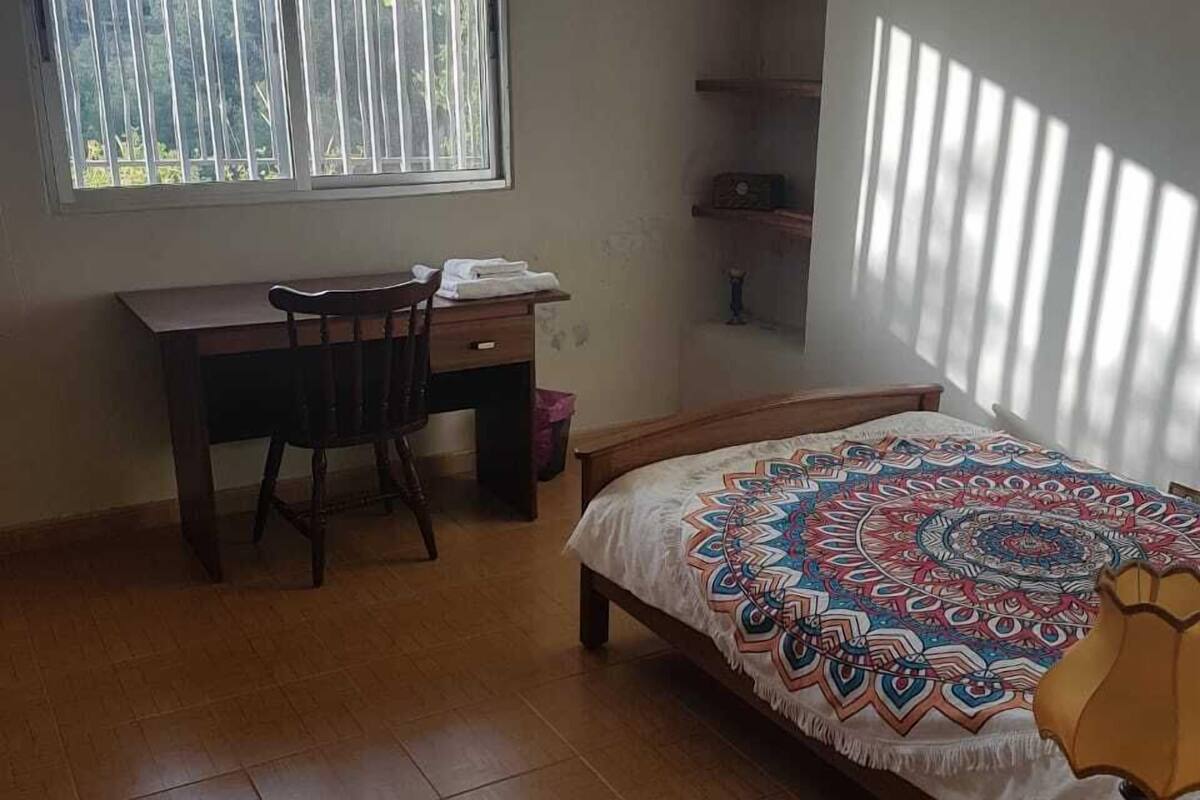 A bedroom with a bed, a desk, and a chair. There is a window with blinds, a shelf, and a lamp.