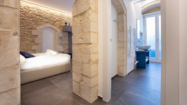 A bedroom with stone walls, a bed, and a view of a bathroom with a large bowl sink. - Ad Maiora - Desing Rooms (Ragusa)