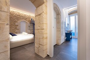 A bedroom with stone walls, a bed, and a view of a bathroom with a large bowl sink. - Ad Maiora - Desing Rooms (Ragusa)