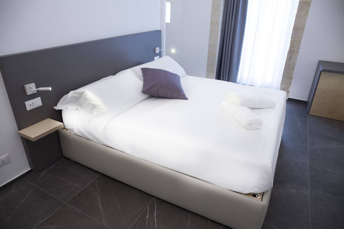 Een net opgemaakt bed met witte lakens en een enkel paars kussen, een nachtkastje met een lamp, en een moderne badkamer zichtbaar door het raam.