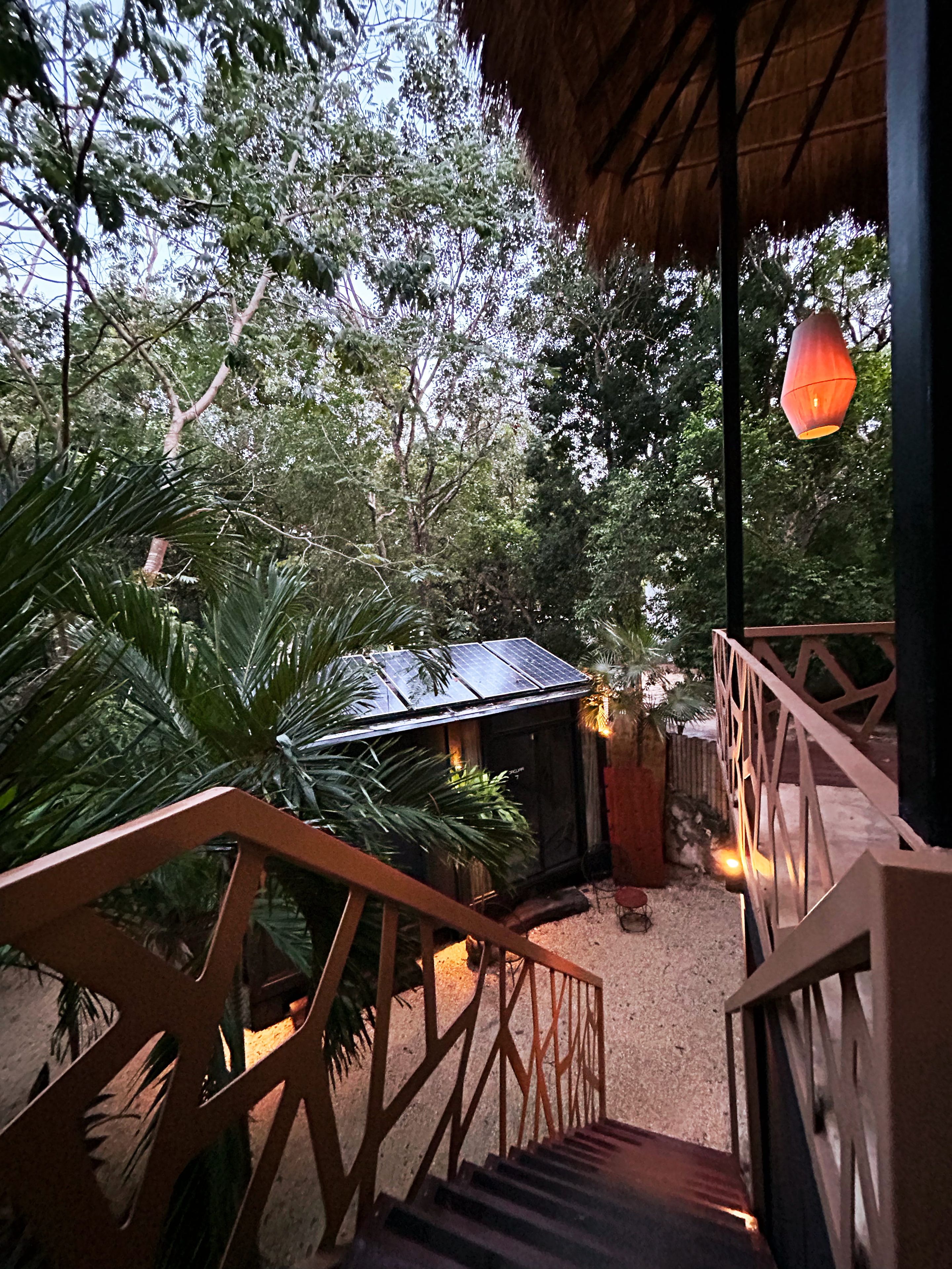 A wooden staircase leading to a covered outdoor area with a thatched roof, surrounded by lush greenery and a small structure with a solar panel.