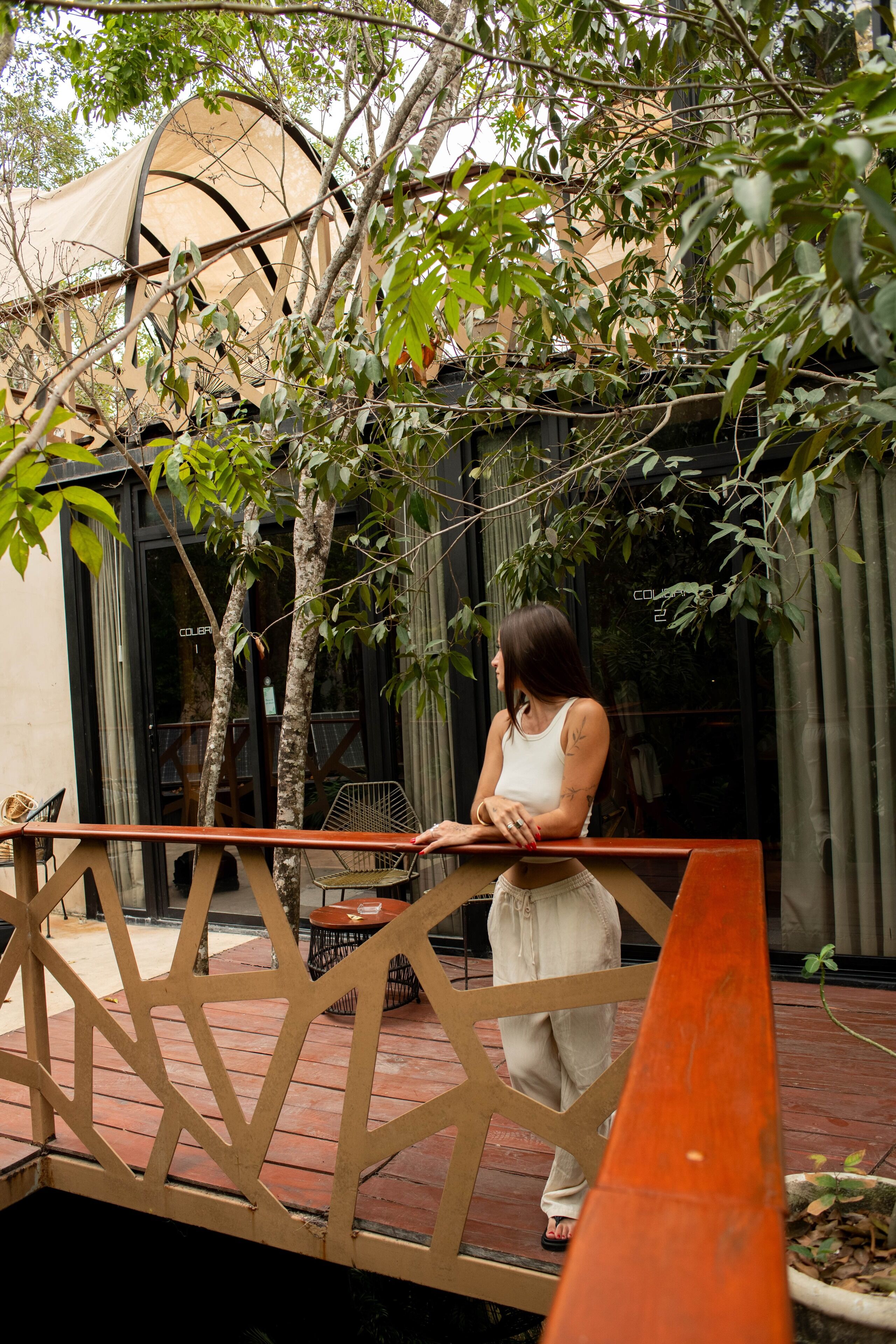Una persona parada sobre una terraza de madera con barandilla de metal, rodeada de árboles y un edificio con ventanas grandes.