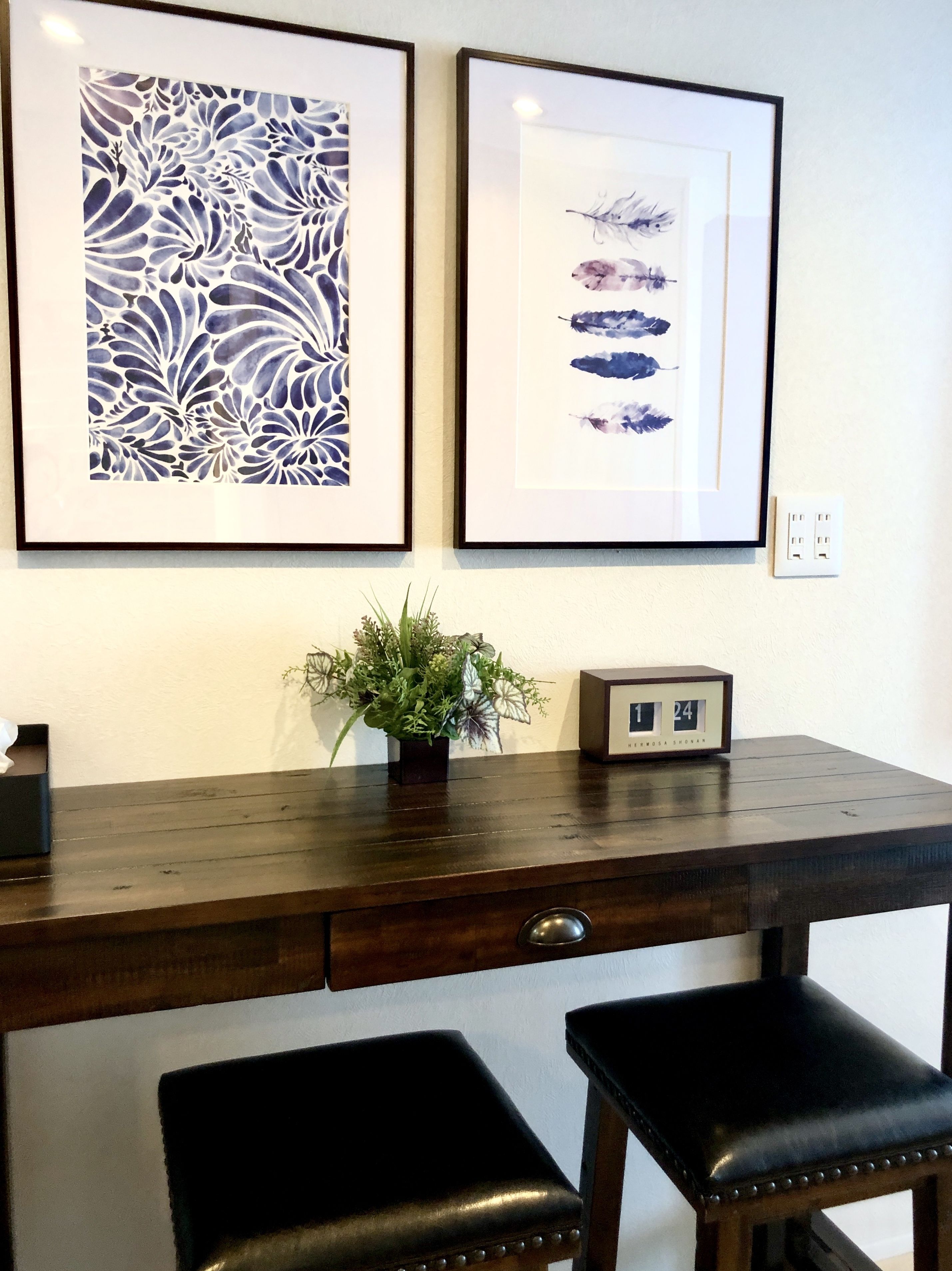 A wooden desk with two black leather chairs, a small potted plant, and two framed artworks on the wall.