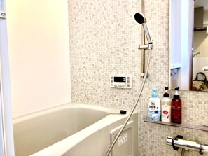 A bathroom with a bathtub, a handheld showerhead, a digital control panel, and toiletries on a shelf.