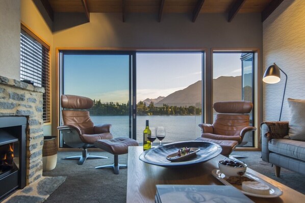 A cozy room with a fireplace, two leather chairs, a dining table with a plate of food, and a large window overlooking a lake and mountains.