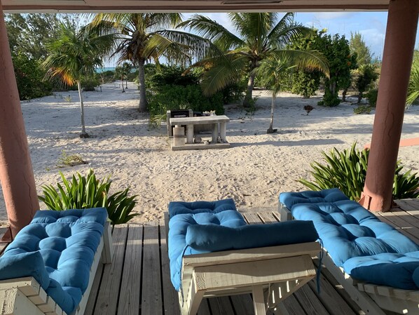 A wooden deck with blue cushioned lounge chairs and a table with chairs, overlooking a sandy beach with palm trees and a clear sky. - 🌴 Sundial Villas ~ Conch Coastal Calm (Bambarra Beach)