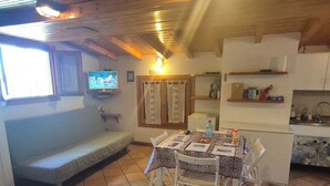 A cozy room with a wooden ceiling, a television on the wall, a bench, a table with chairs, and a kitchen area with shelves. - Lakeside cottage with private garden and pier. Pet-friendly.  (Oliveto Lario)