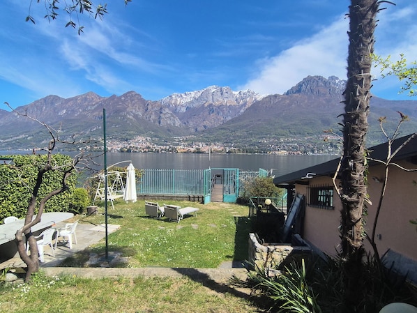 Vista del lago con montañas al fondo, un patio con sillas, un conjunto de columpios y un edificio pequeño con una puerta.