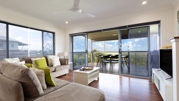 A modern living room with a sofa, coffee table, and a dining area with chairs. There is a large sliding glass door leading to an outdoor deck with a view of the landscape. - Jinalong - Breathtaking Views (Angourie)