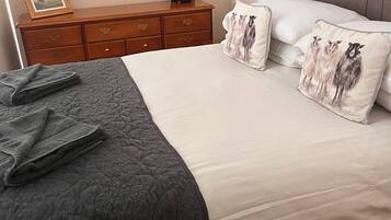 Una cama bien hecha con colcha gris y ropa de cama blanca, un aparador de madera con un espejo, y un cuadro en el aparador.