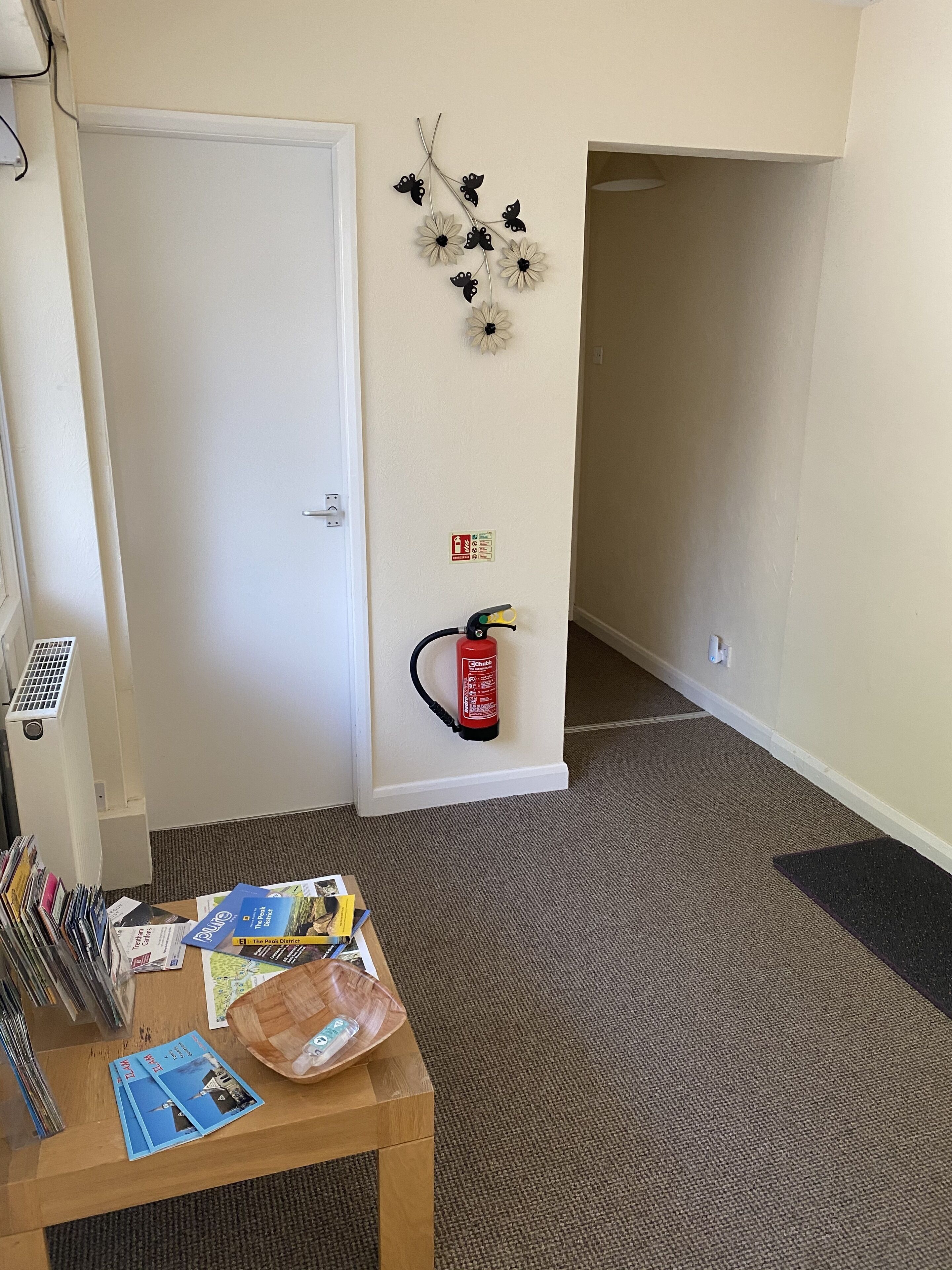 Ein Zimmer mit geschlossener Tür, einem an der Wand montierten Feuerlöscher, einer mit Papieren besetzten Schwarzwerttafel und einem Tisch mit Zeitschriften und Tablett.