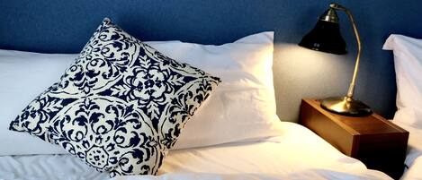 A neatly made bed with white linens and a single decorative pillow with a blue and white pattern. A black desk lamp is positioned on a wooden nightstand beside the bed.