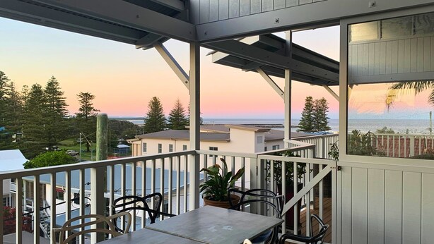Un balcón con mesa y sillas, con vista a una zona residencial y un cuerpo de agua al atardecer.