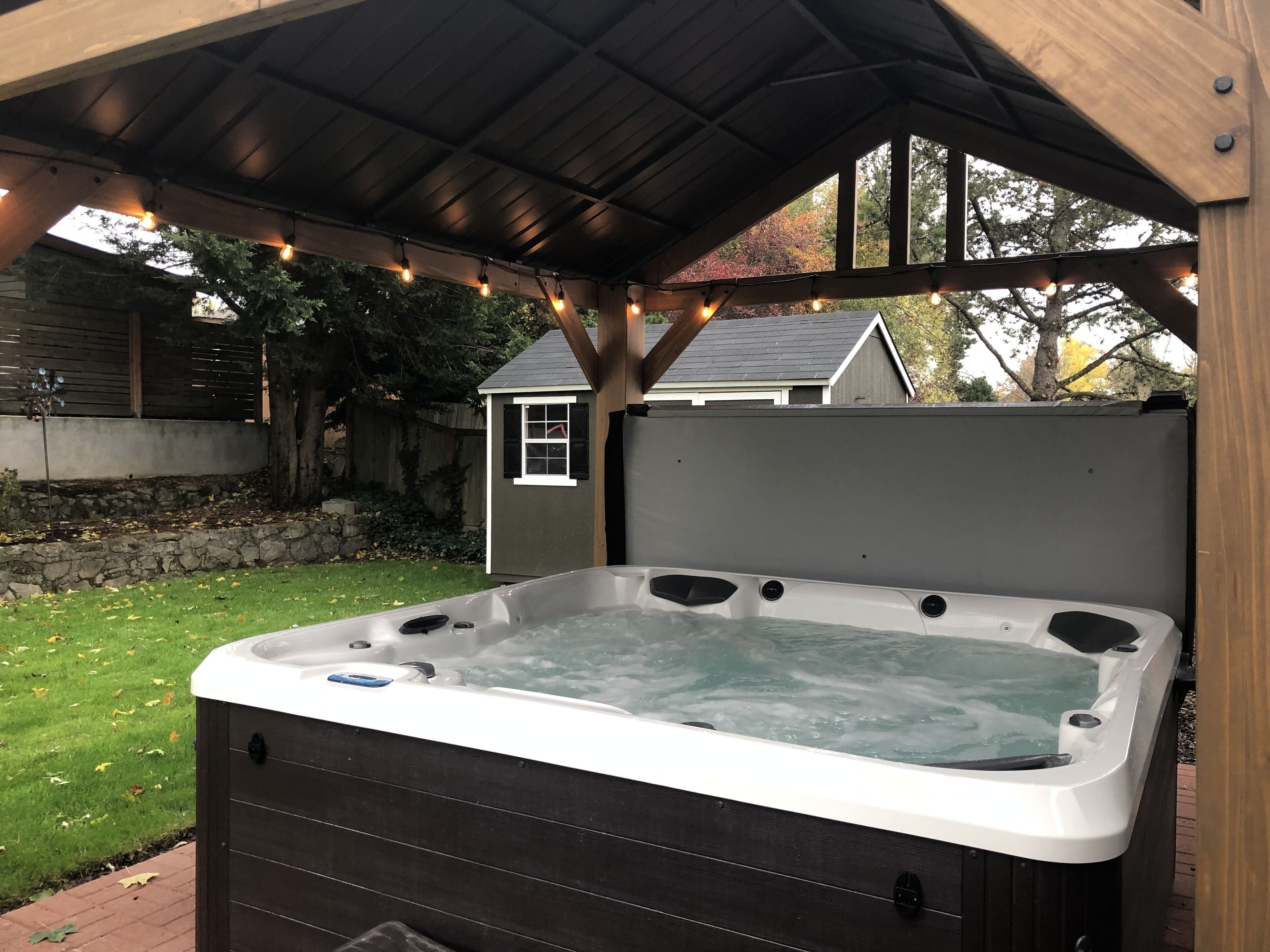 A hot tub under a wooden pergola with string lights, surrounded by a grassy yard and a stone wall.