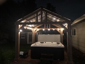 Un jacuzzi bajo un porche de madera con luces de cadena, rodeado por un jardín.