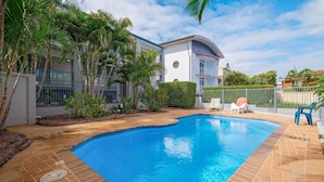 Une piscine dont le bord est revêtu de carreaux, entourée d’un jardin aménagé avec des palmiers et un bâtiment blanc doté d’un toit incurvé.