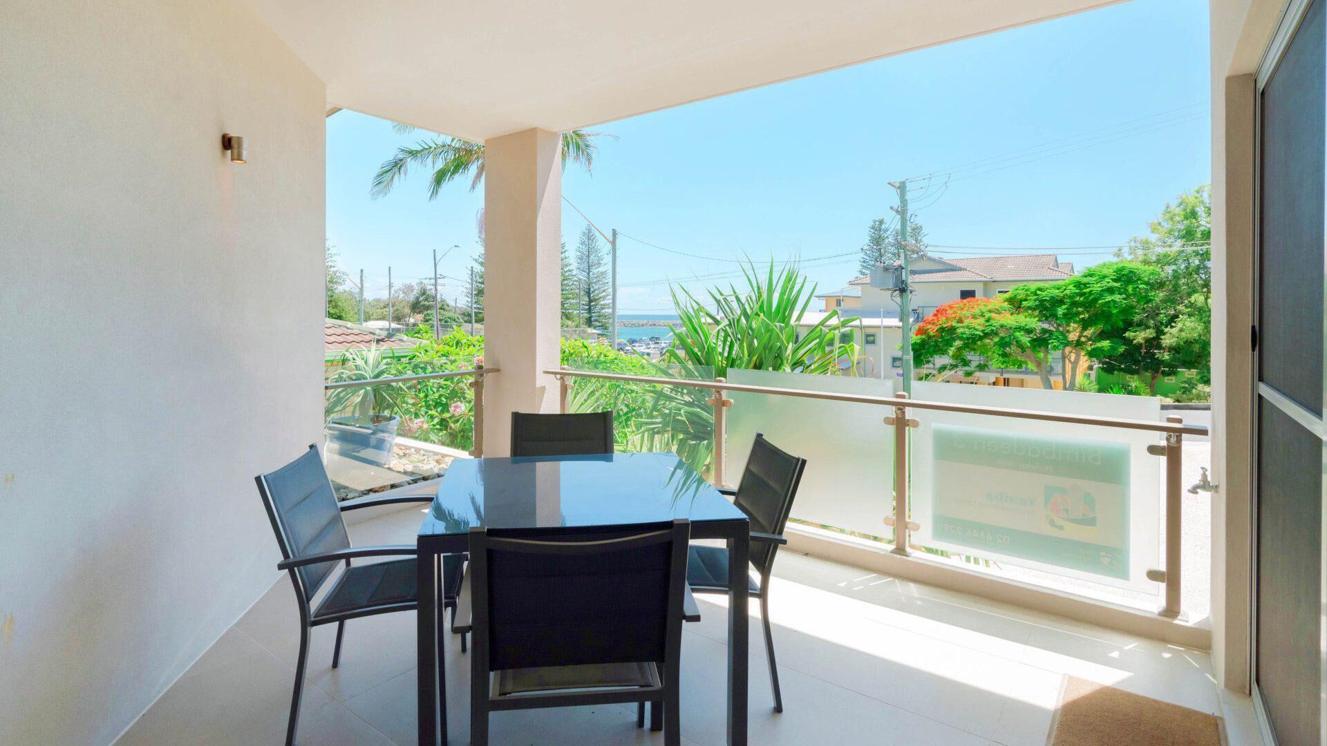 A balcony with a table and chairs, offering a view of the sea and surrounding greenery.