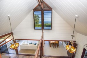 Ein Loft-Zimmer mit hohen Decken, Holzbalken, einem weißen Sofa, einem Esstisch und einem großen Fenster mit Blick.