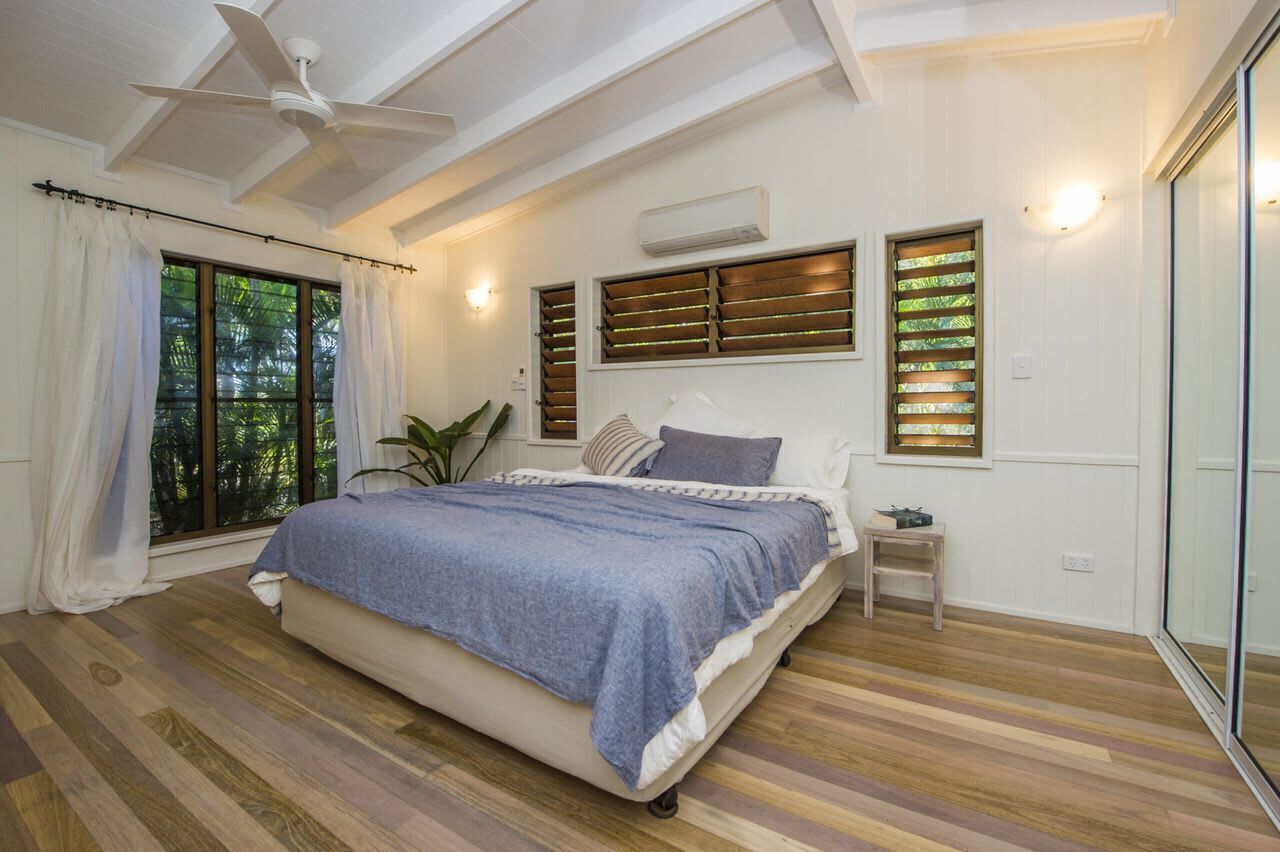 A bedroom with a bed, ceiling fan, large windows with blinds, and a sliding glass door.