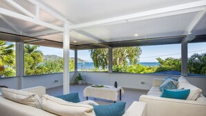 Ein moderner Außenbereich mit einer weißen Couch, einem kleinen Tisch und Blick auf das Meer und viel Grün.
