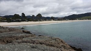 A coastal landscape with a rocky foreground, a sandy beach, and a backdrop of hills and trees under a cloudy sky. - Private stunning elevated ocean veiws (Whangapoua)