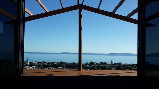 A room with a large window offering a view of a coastal landscape. - Private stunning elevated ocean veiws (Whangapoua)
