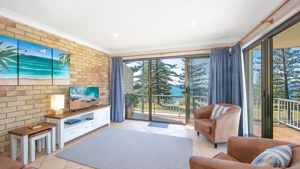 A spacious living room with a television, a sofa, and a chair, a brick wall, and a large sliding glass door leading to a balcony with a view of trees and a body of water. - Flinders Lodge - an idyllic holiday rental (Yamba)
