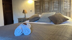 Ein ordentlich bezogenes Bett mit aufgerollten Handtüchern, einer Lampe und Holzjalousien an der Wand.