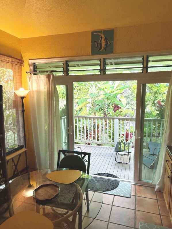 A room with a sliding glass door leading to a balcony with a view of greenery. There is a table with chairs and a lamp. - Quiet, Licensed, Clean & Affordable Cottage Studio with Kitchenette (Princeville)