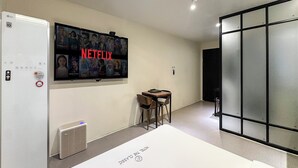 Une chambre d’hôtel avec un grand téléviseur fixé au mur, sur lequel est affichée une affiche de Netflix, une petite table avec des chaises, un radiateur et une porte-fenêtre coulissante.