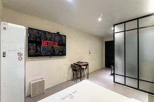 Habitación de hotel con un televisor de gran tamaño montado en la pared mostrando un cartel de Netflix, una mesita con sillas, un radiador y una puerta de vidrio corredera.