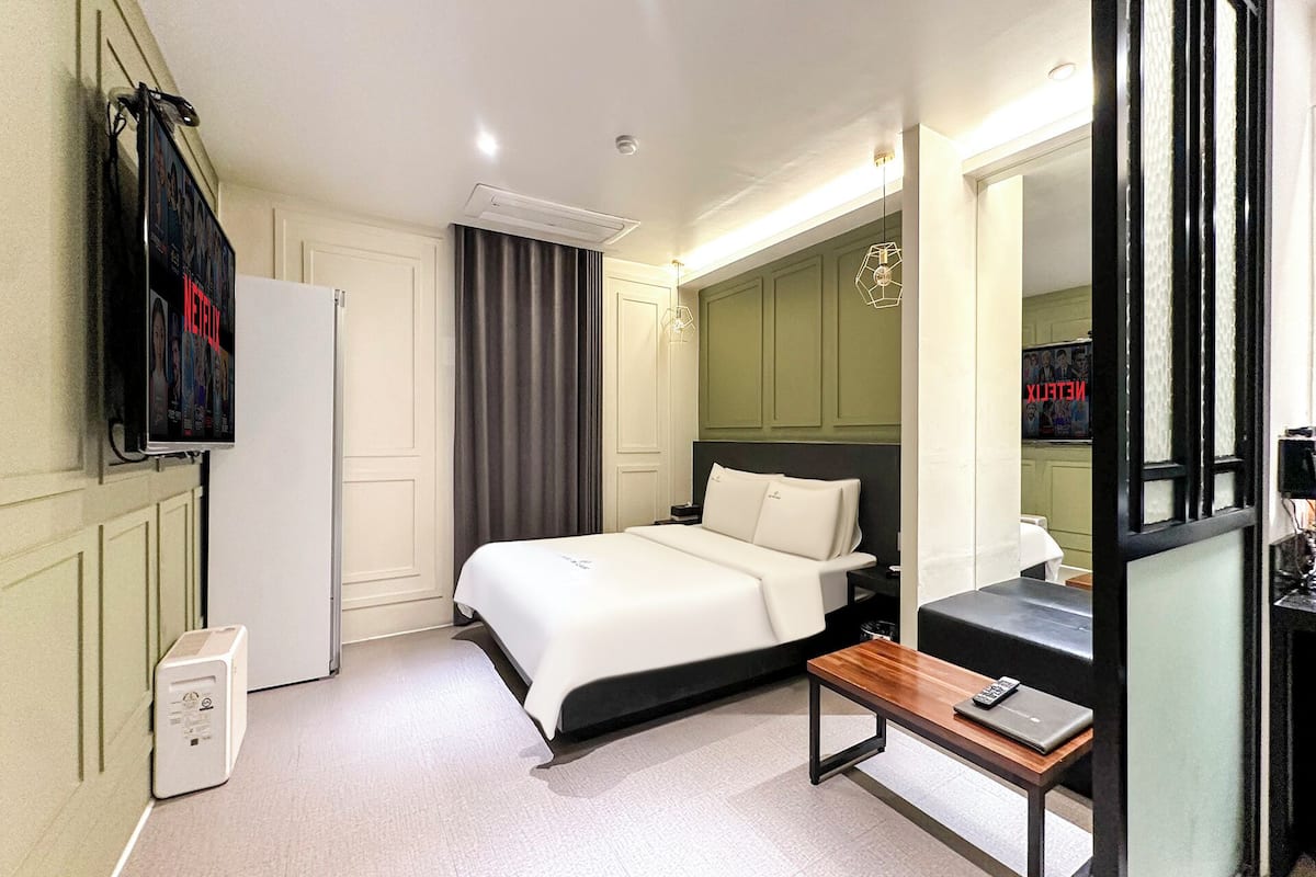 Quarto de hotel com cama, mesa de cabeceira, escrivaninha com cadeira, televisão fixada na parede e janela com cortinas.