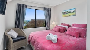 Une chambre à coucher avec un grand lit, une chaise en osier, une fenêtre avec des rideaux et un tableau accroché au mur.