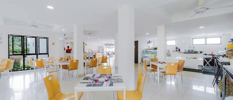 Un vaste espace dédié à la salle à manger, avec des tables blanches et des chaises jaunes, une cuisine en arrière-plan et de grandes fenêtres laissant entrer la lumière naturelle.
