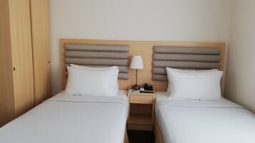 A hotel room with two single beds, a wooden wardrobe, a bedside table with a lamp, and a nightstand with a phone and a small box.