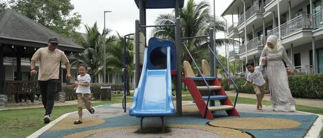 Un parque infantil con un tobogán, rodeado de una zona ajardinada con palmeras y un edificio al fondo.