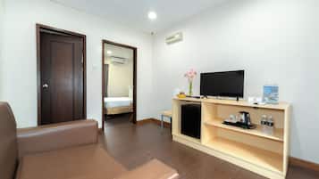 A modern hotel room with a brown leather sofa, a TV on a wooden stand, a small table with a vase of flowers, and a bed in the background.