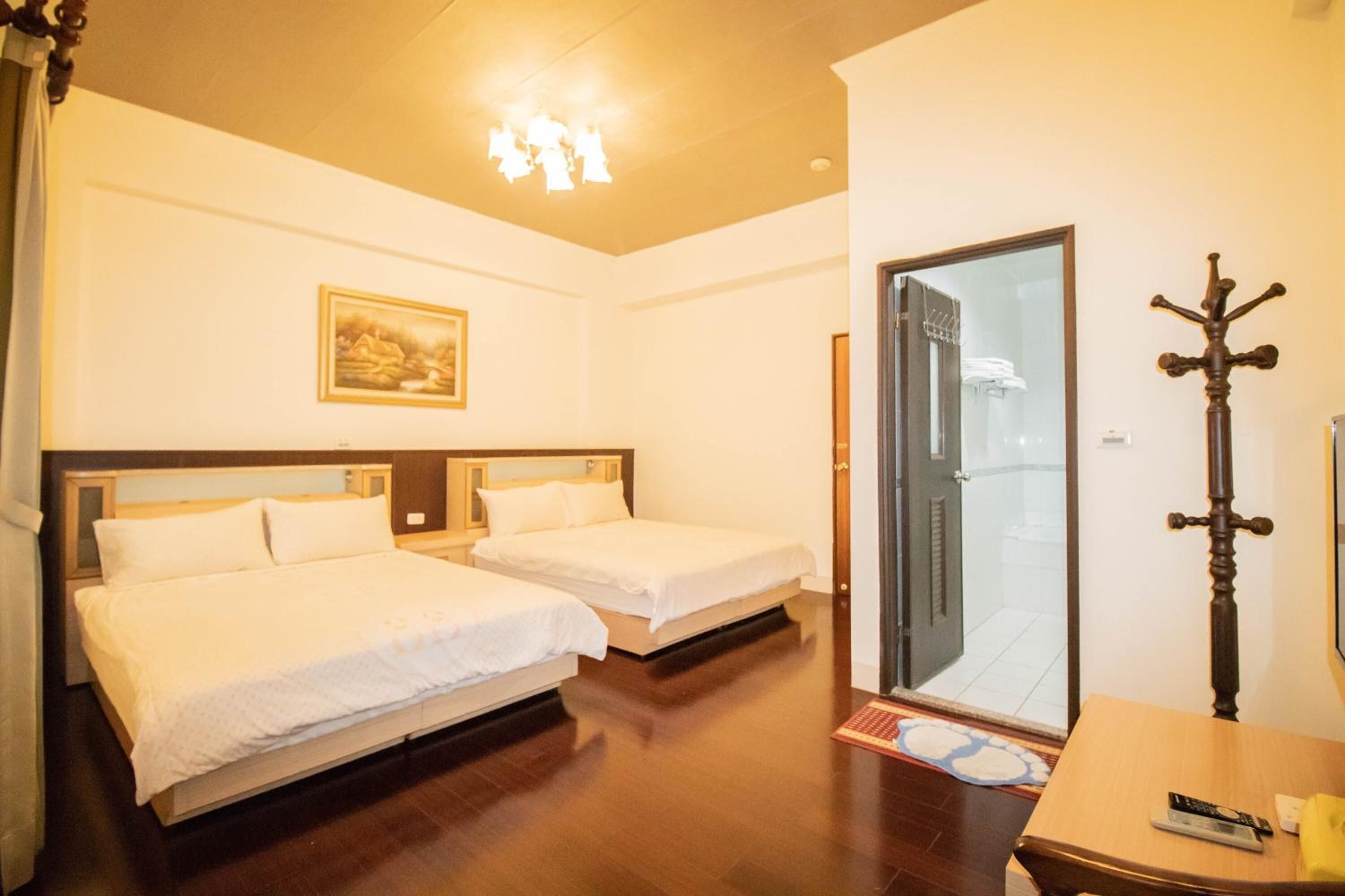 A hotel room with two beds, a wooden floor, a painting on the wall, a coat rack, and a door leading to a balcony.