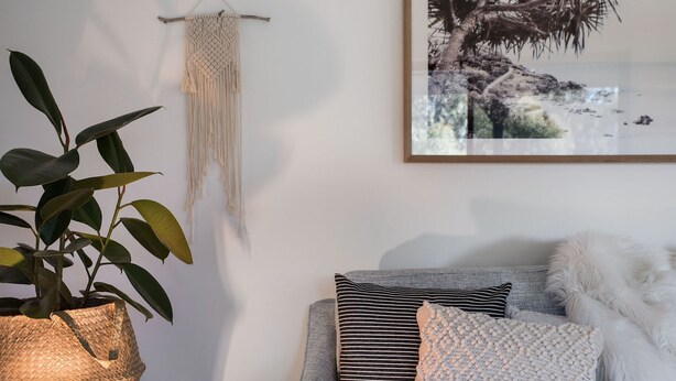 Un salon agrémenté d’une plante en pot tressée, d’une décoration murale en macramé et d’un tableau encadré représentant un paysage côtier.