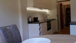 Una cocina compacta con cafetera, hervidor y grifo moderno. En primer plano, una mesa de comedor con un mantel individual.