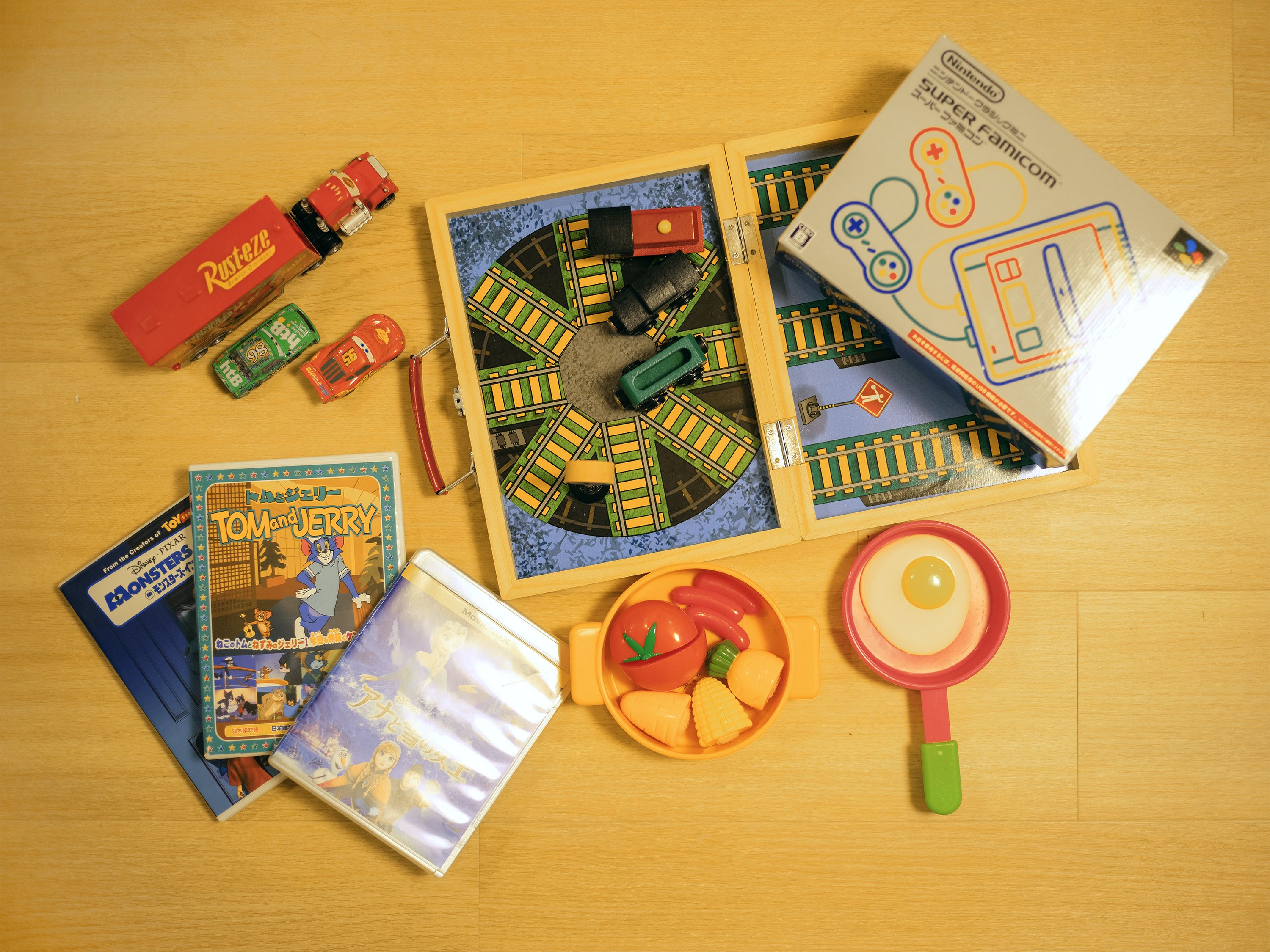 A wooden table with a train set, a toy car, a toy magnifying glass, a toy bowl with vegetables, and a few books.